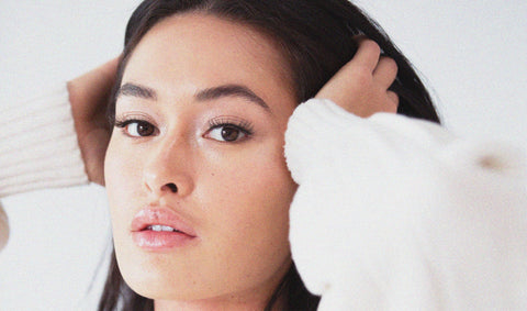 light skinned brunette model with freckles and both hands combing through long dark hair, wearing false Lithe Lashes, wearing white sweater in light grey background