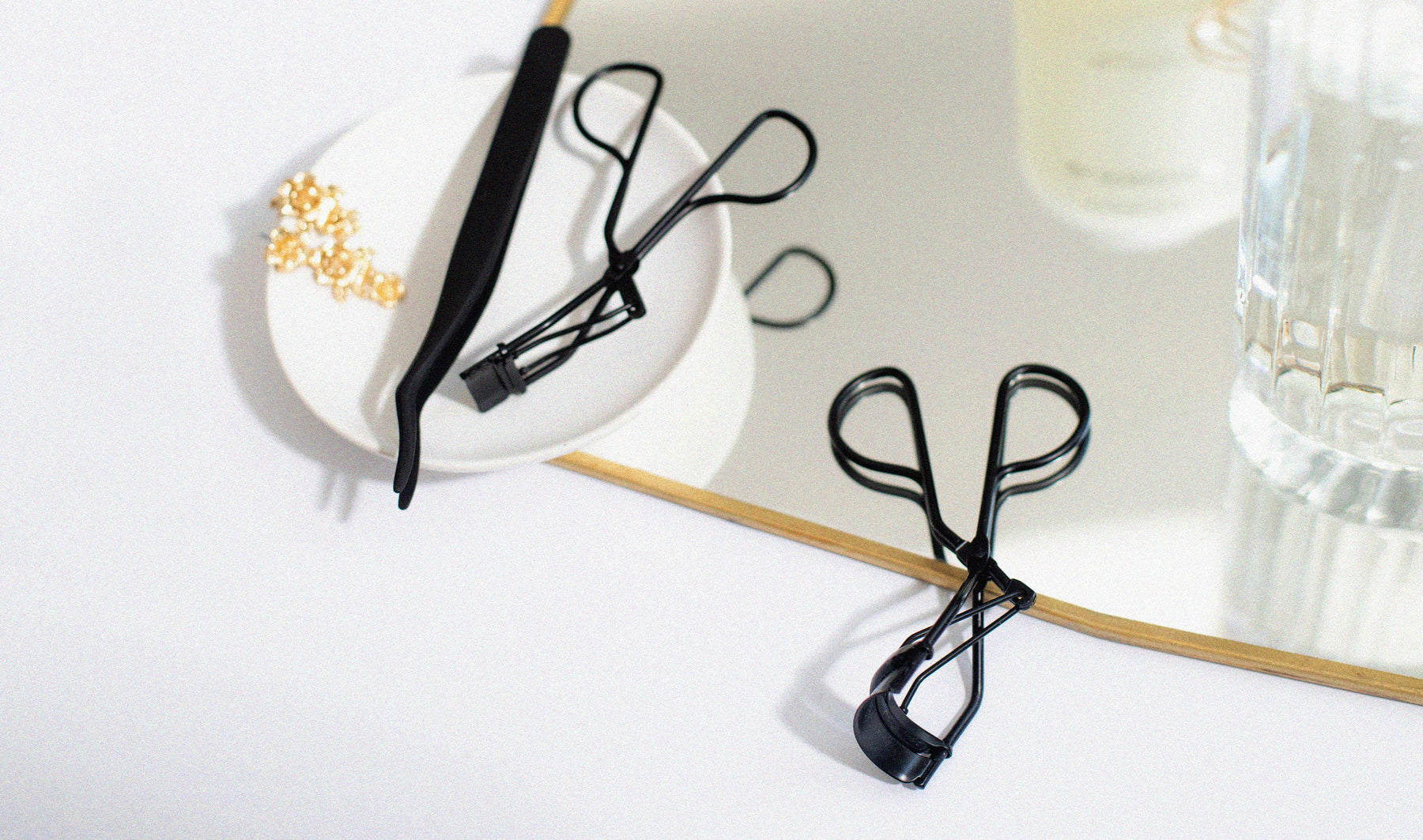 Matte black Lithe Lash Applicator on a stylish plate, next to black Lithe Mini Lash Curler and Lithe Lash Curler on a white and light grey back ground, resting partially on a stylish clear and clean mirror 