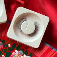 Lithe Lashes Mini Lashes lash style named M1 - Mini Round, a product image of the lashes in their biodegradable stowing tray, laying overtop red floral silk fabric.