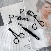 Lithe Lashes false lashes accessory, the Lithe Lash Scissors. A product image showing all Lithe's matte black lash tools, with the Lash Scissors in the bottom left, the Mini Lash Curler at the top, the Lash Applicator at the bottom of the page, and the Lithe Lash Curler in the upper right portion of the image, all of which are laying gently on black and white magazine print.