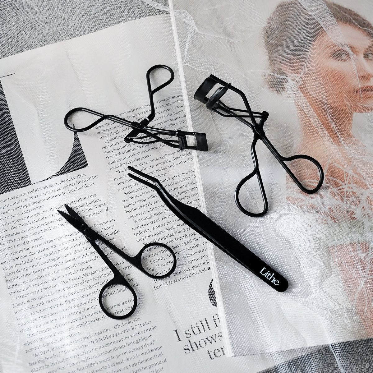 Lithe Lashes false lashes accessory, the Lithe Mini Lash Curler. A product image showing all Lithe's matte black lash tools, with the Lash Scissors in the bottom left, the Mini Lash Curler at the top, the Lash Applicator at the bottom of the page, and the Lithe Lash Curler in the upper right portion of the image, all of which are laying gently on black and white magazine print.
