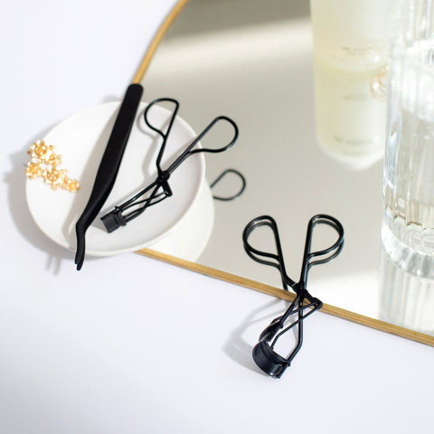 Lithe Lashes false lashes accessory, the Lithe Lash Curler. A product image showing the matte black Lash Applicator and the Mini Lash Curler resting on a small white porcelain plate, while the ergonomic Lithe Lash Curler is resting half on a mirror with thin golden frame and the other half on a spotless white table. In the background is an expensive bottle of perfume and crystal candle holder.